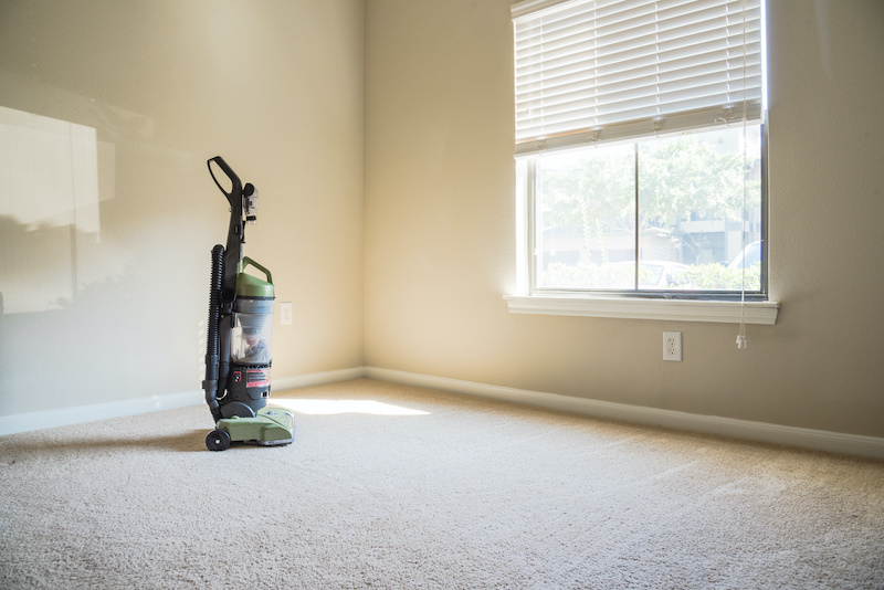 Modern vacuum cleaner stands at the corner of clean apartment bedroom with window view, natural light and vacuuming rough carpet. Typical apartment bedroom detail in America.